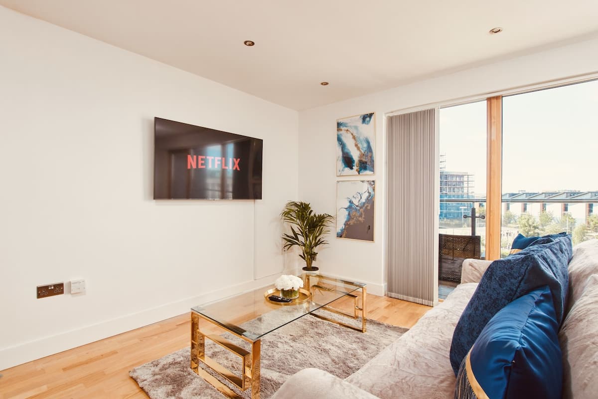 Ein modernes Wohnzimmer mit einem Glastisch, einer blauen Sofaausstattung und einem großen Fenster mit Blick auf Gebäude. Eine an der Wand befestigte TV-Gerät zeigt 'NETFLIX'.
