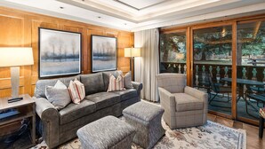 Ein Wohnzimmer mit Sofa, Hocker und Ohrensessel, einem Couchtisch, einem Fernseher und einem großen Fenster mit Blick ins Freie.