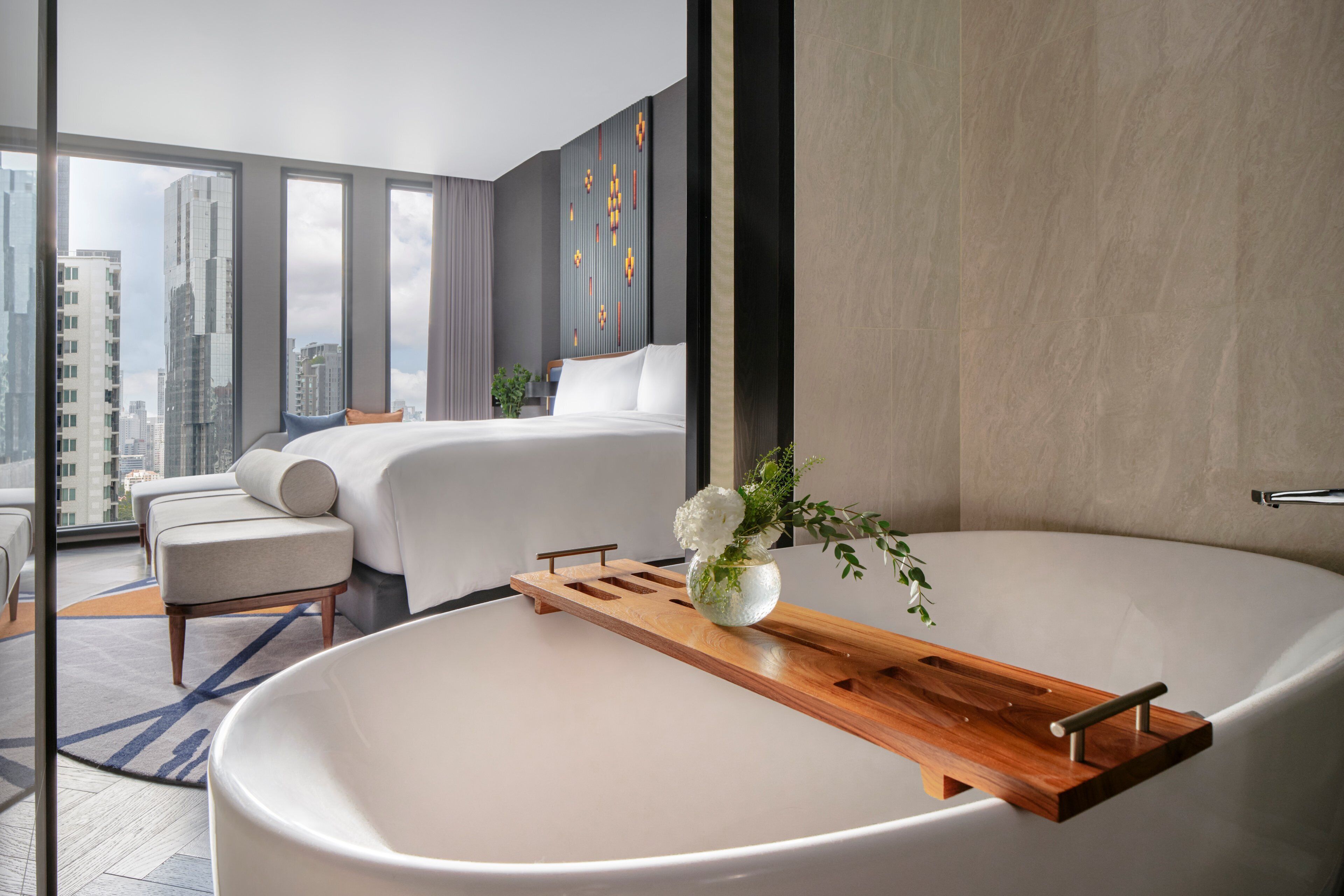 A modern hotel room with a large bathtub, a bed with white linens, a wooden tray, and a vase with flowers.
