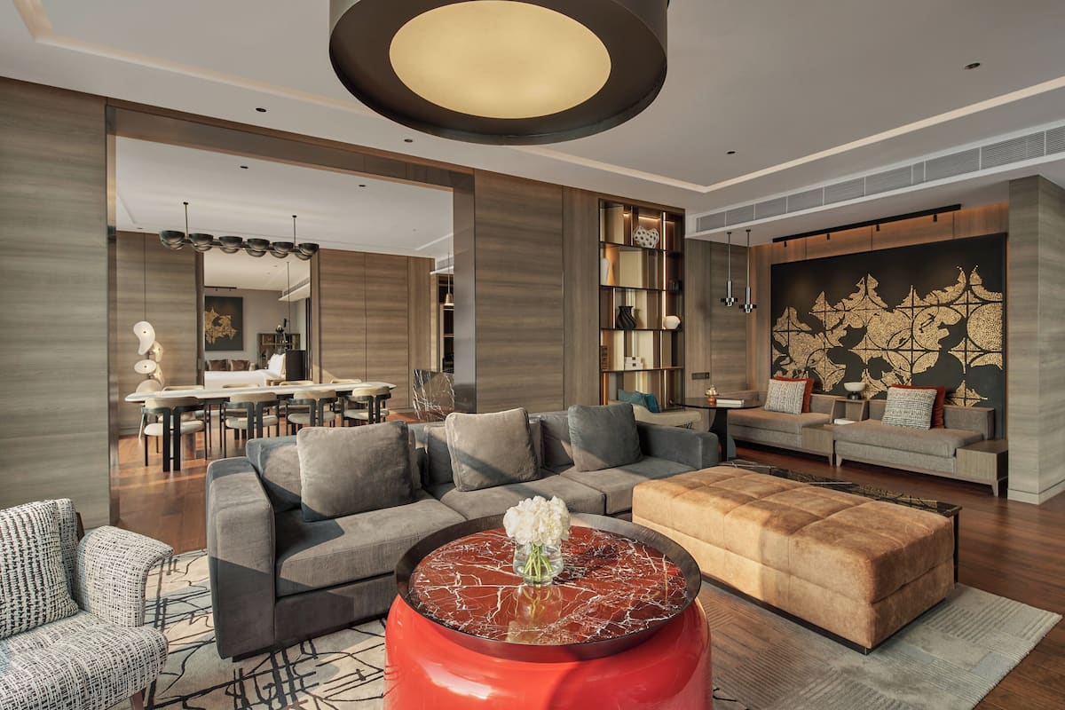 A modern living room with a sectional sofa, a red ottoman, and a dining area in the background.