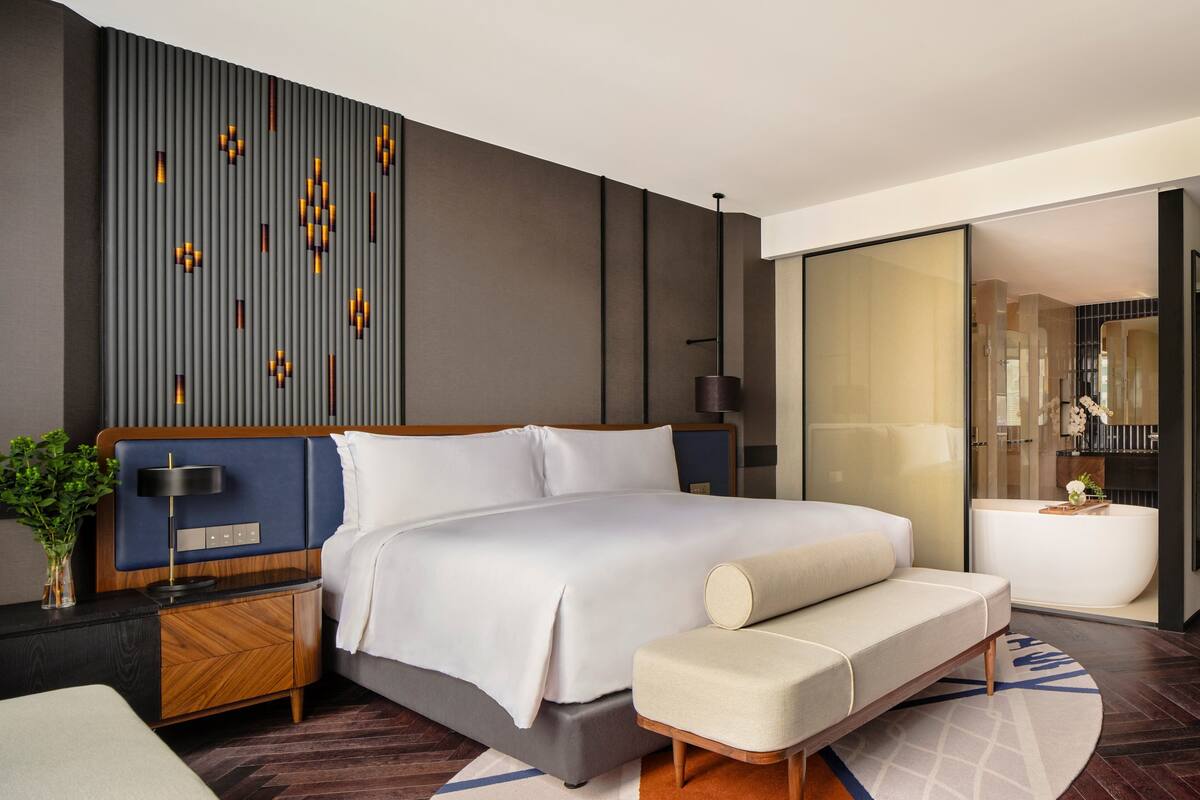 A modern hotel room with a large bed, a bedside table with a lamp, a bench, and a view of a bathroom with a bathtub.