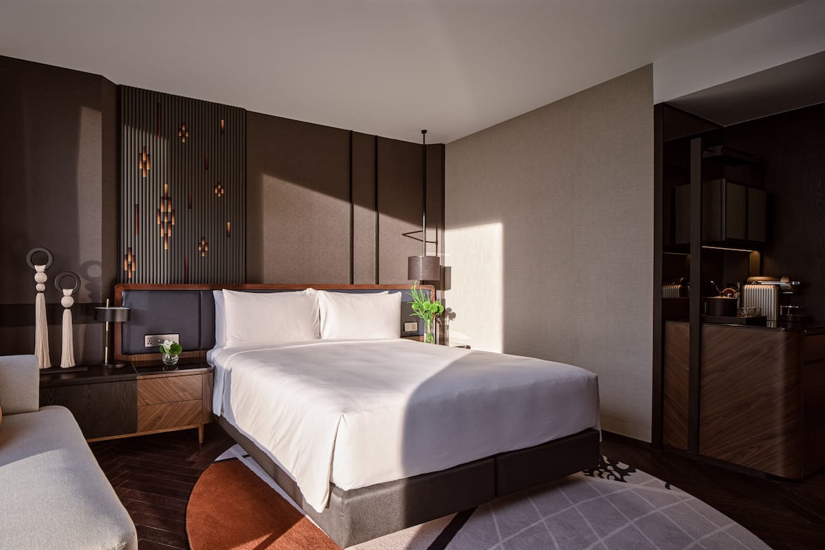 A modern hotel room with a large bed, a sitting area with a sofa and armchair, a nightstand with a lamp, and a view of the city through large windows.