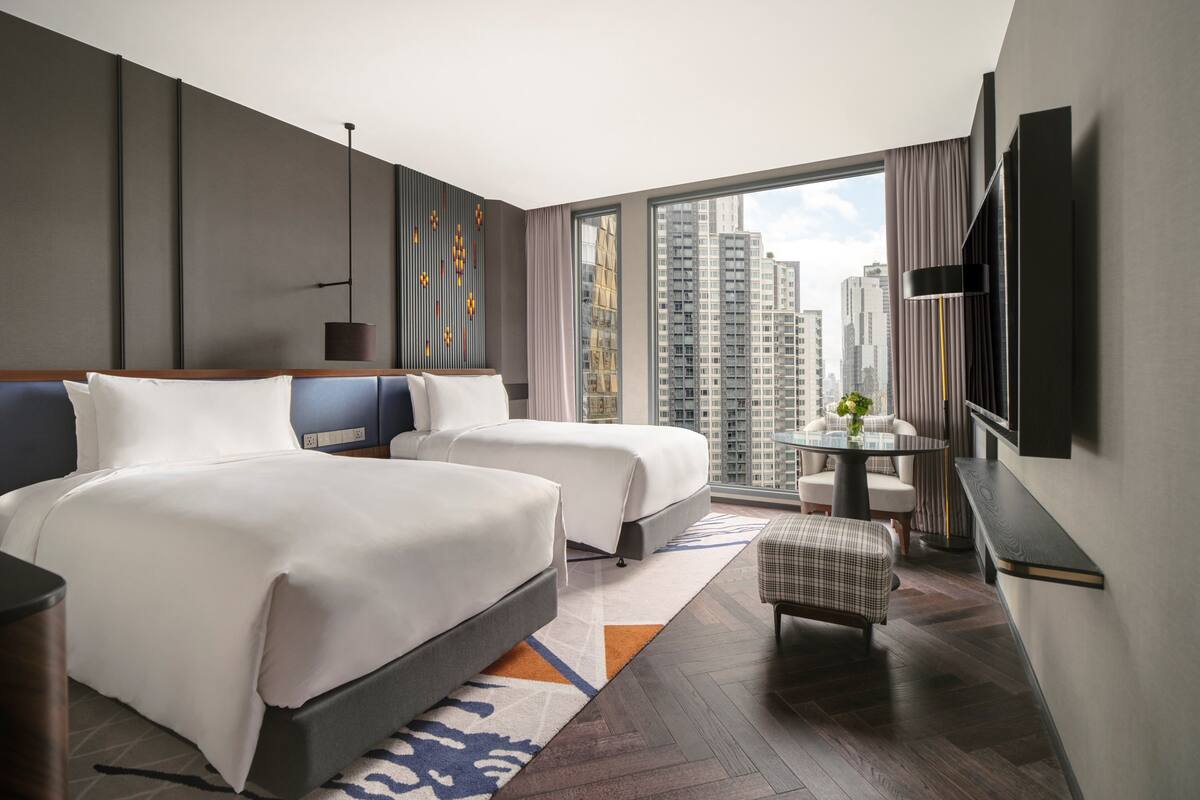 A modern hotel room with two beds, a small table with a vase, a chair, and a large window offering a city view.