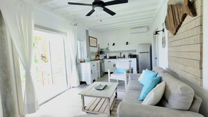 A bright, open-plan living area with a ceiling fan, a sofa with cushions, a coffee table, and a view of the kitchen in the background.