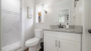 A bathroom with a white toilet, a shower, a vanity with a mirror, and a framed quote on the wall.