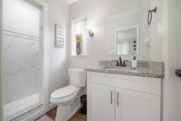 A bathroom with a white toilet, a shower, a vanity with a mirror, and a framed quote on the wall.