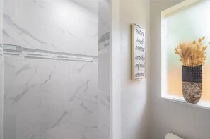 Une salle de bain avec un mur en marbre, une citation inspirante encadrée et une fenêtre ornée d’un vase de fleurs séchées.