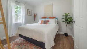 A bedroom with a bed, a window with curtains, a plant, and a wooden floor.