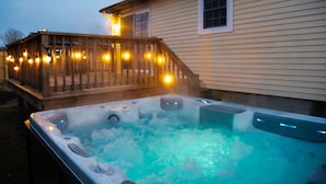 A hot tub with a wooden deck and string lights, adjacent to a house with a window and a door. - NEW! Updated Mystic Home w/ Sauna, Hot Tub & Deck (Groton)