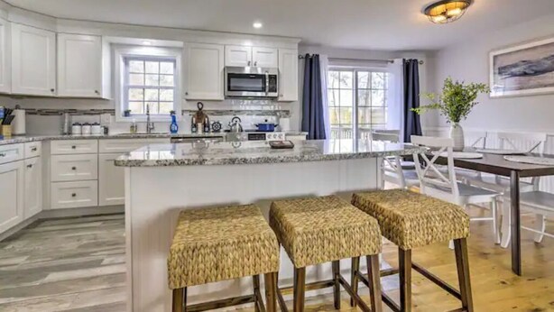 A modern kitchen with white cabinets, a central island with stools, and a dining area with a table and chairs. - NEW! Updated Mystic Home w/ Sauna, Hot Tub & Deck (Groton)