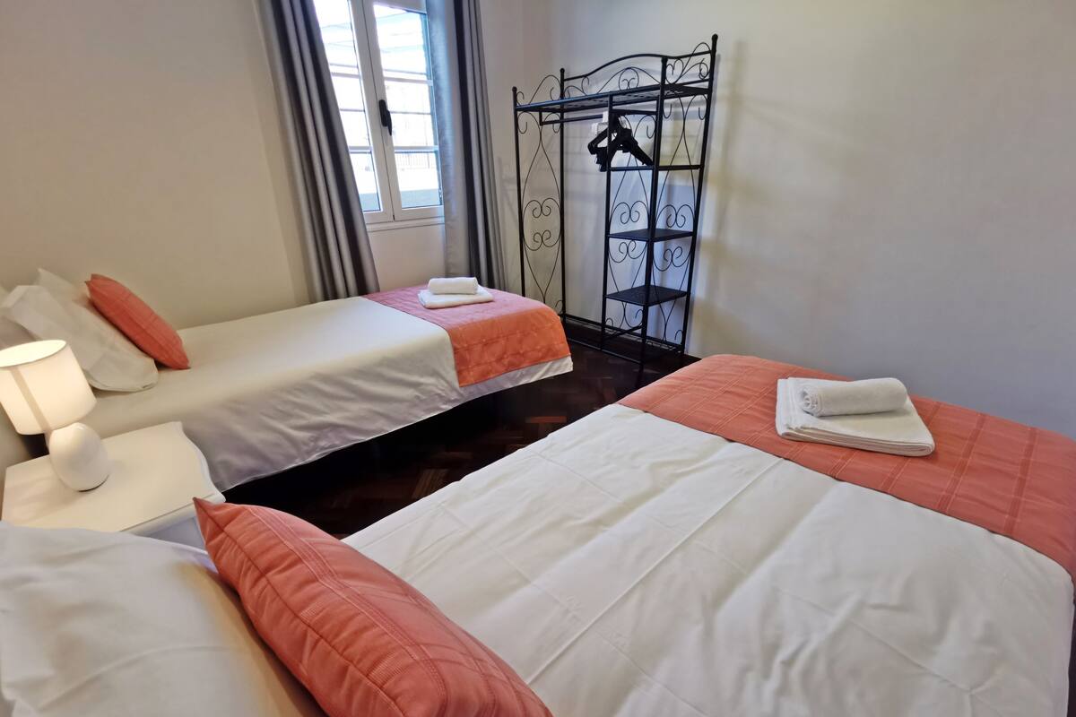 A hotel room with two single beds, each with a white and orange bedspread, a nightstand with a lamp, and a metal rack with clothes hanging.