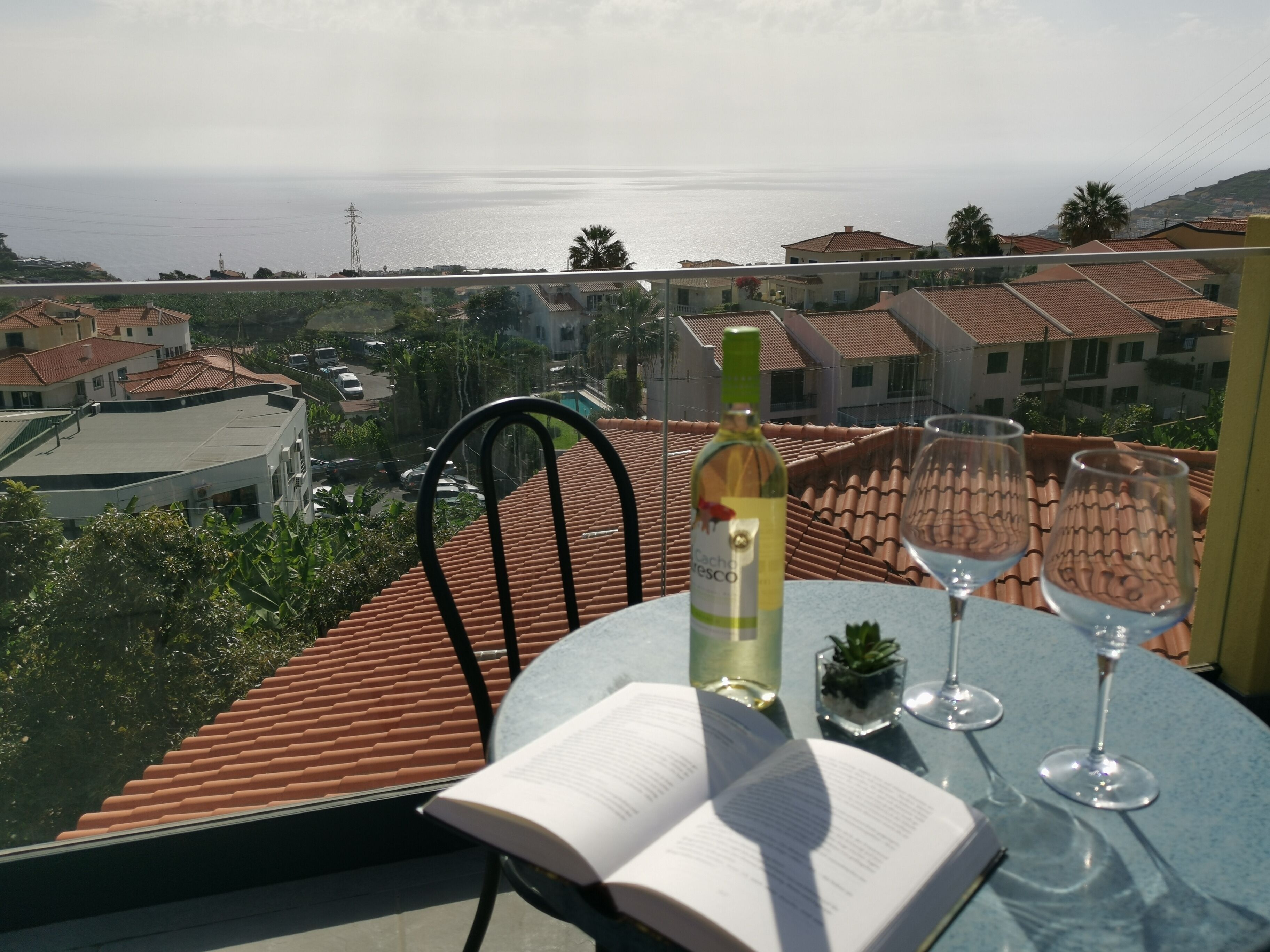 A balcony with a view of the sea, a bottle of wine, two wine glasses, and an open book.