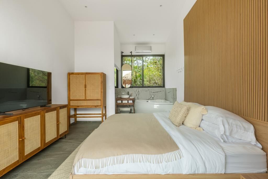 Une chambre moderne avec un grand lit, une armoire en bois, une télévision, une fenêtre donnant sur un paysage verdoyant, et une salle de bain avec une baignoire et un lavabo.