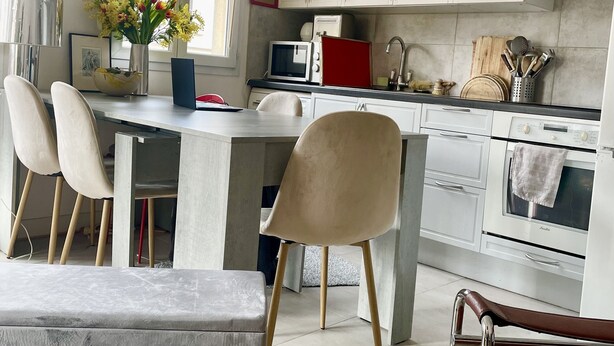 Una cocina moderna con armarios blancos, un microondas integrado y un horno. Hay un comedor con un banco y sillas, y un florero con flores en la encimera.