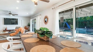 A modern dining area with a wooden table, white chairs, and woven placemats. - Luxury Tropical Modern Pool Estate/NO PARTIES (San Bernardino)