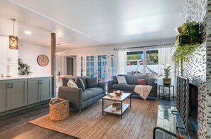 Uma sala de estar moderna com sofá, mesa de centro e lareira. Há uma área de cozinha com armários e um relógio na parede.