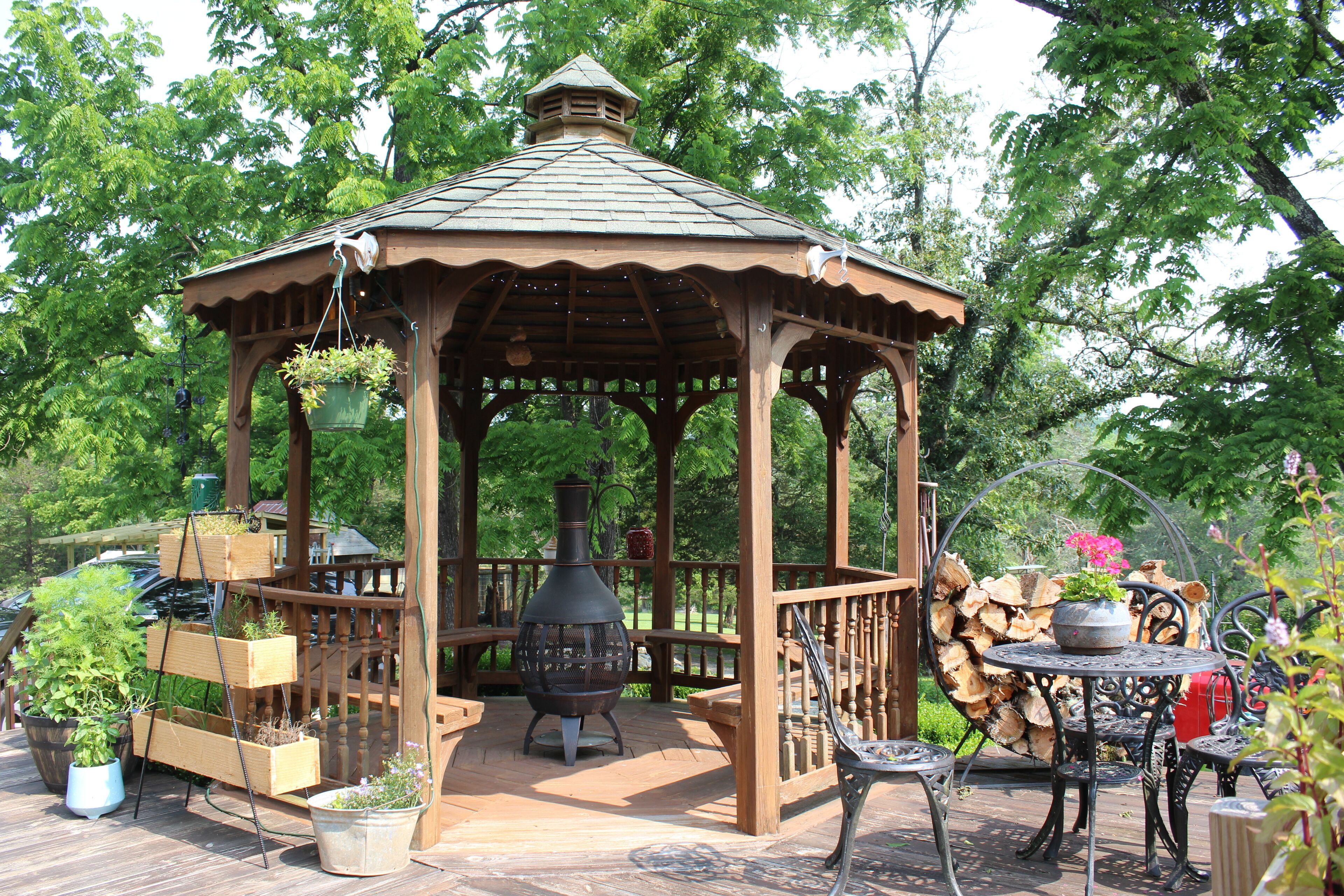 A wooden gazebo with a tiled roof, surrounded by potted plants and a wood stove.