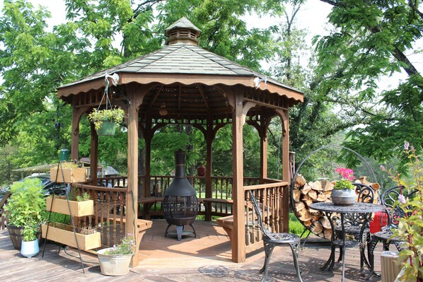 A wooden gazebo with a tiled roof, surrounded by potted plants and a wood stove.