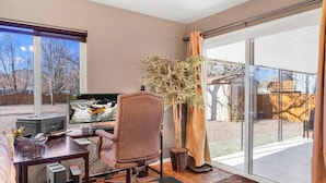 Ein Home-Office mit Schreibtisch, Stuhl, Computer und Fernseher. Ein großes Fenster bietet Blick auf einen Garten.
