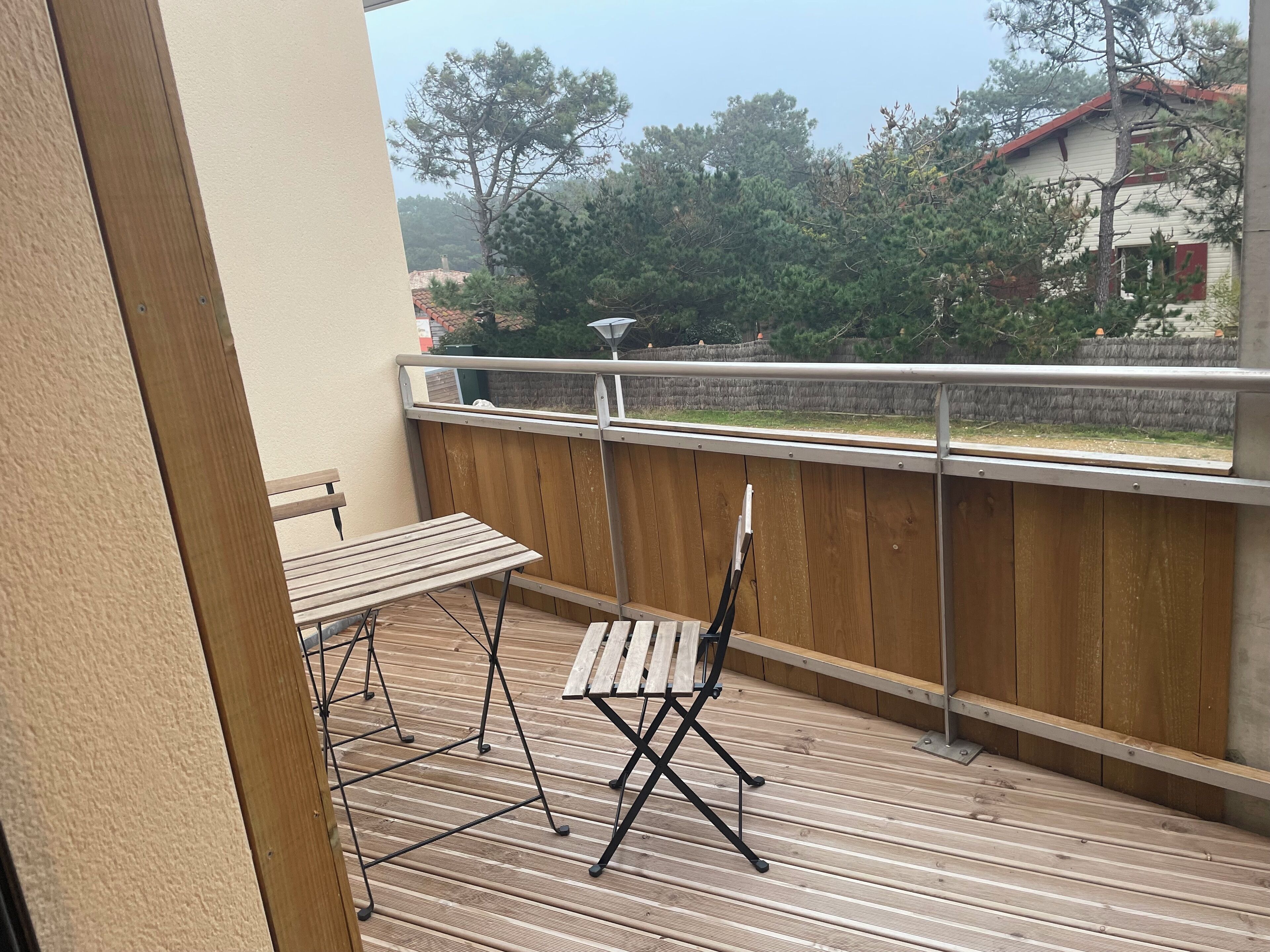 A balcony with a wooden floor, a metal railing, and a view of trees and buildings.