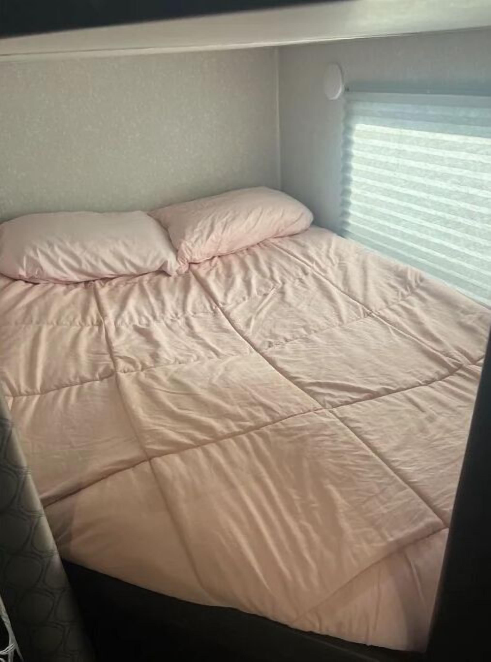A bed with a quilted cover and pillows, a window with blinds, and a partial view of a curtain.
