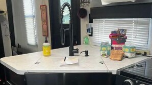 Une cuisine avec un plan de travail noir, un évier, une cuisinière et des placards. On y trouve une fenêtre avec des stores, une horloge et une bouteille de liquide vaisselle sur le plan de travail.