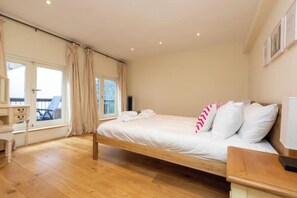 Une chambre avec un lit en bois, un bureau et une chaise. Il y a une fenêtre avec des rideaux, et on aperçoit un balcon à l’extérieur.