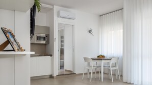 A modern kitchen with white cabinets, a microwave, and an air conditioning unit. There is a dining area with a table and chairs, and a view of a balcony with white curtains. - Harmony House (Piano di Sorrento)