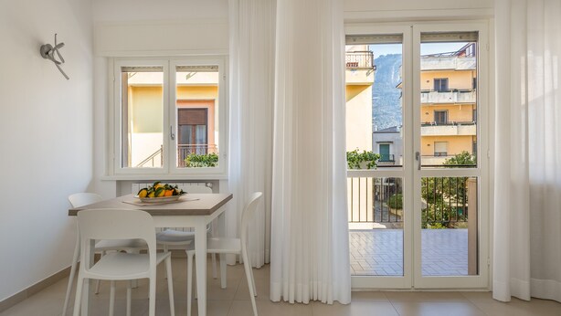A bright, modern apartment with a dining area, white furniture, and a view of neighboring buildings through large windows. - Harmony House (Piano di Sorrento)