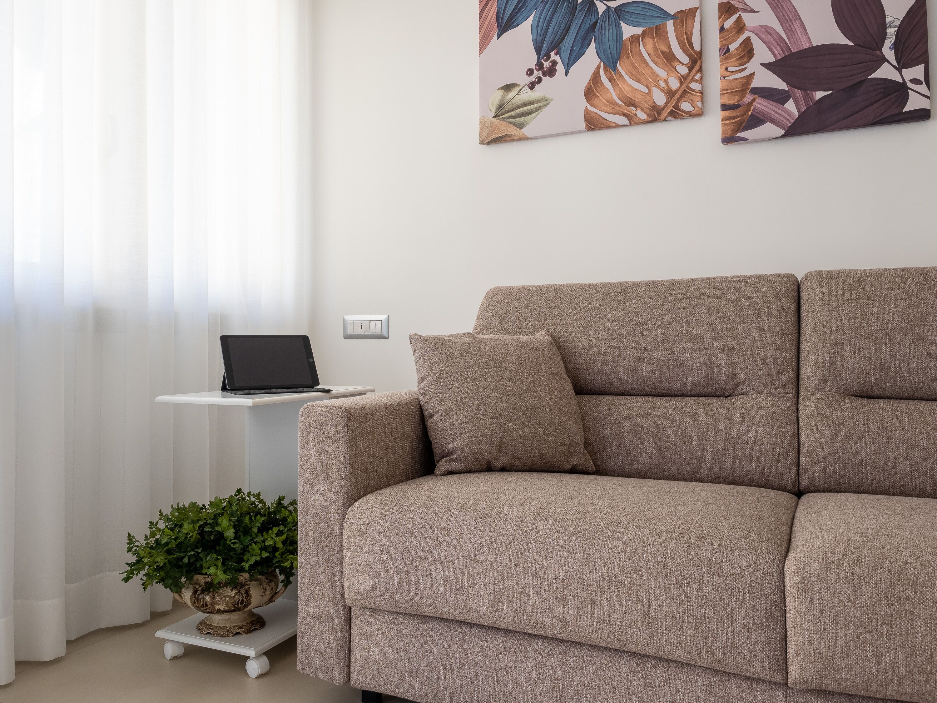 Ein Wohnzimmer mit einem beige-farbenen Sofa, einem kleinen weißen Beistelltisch mit einem Laptop, einer Zimmerpflanze und einem wandmontierten Lichtschalter.