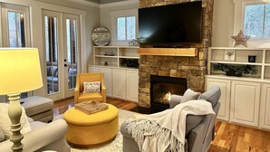 A living room with a stone fireplace, a flat-screen TV, and a comfortable seating area. - Buford Forest Retreat- Fire Pit - 5 bdrms- Sleeps 13 - Water Park, Venues, Golf! (Buford)