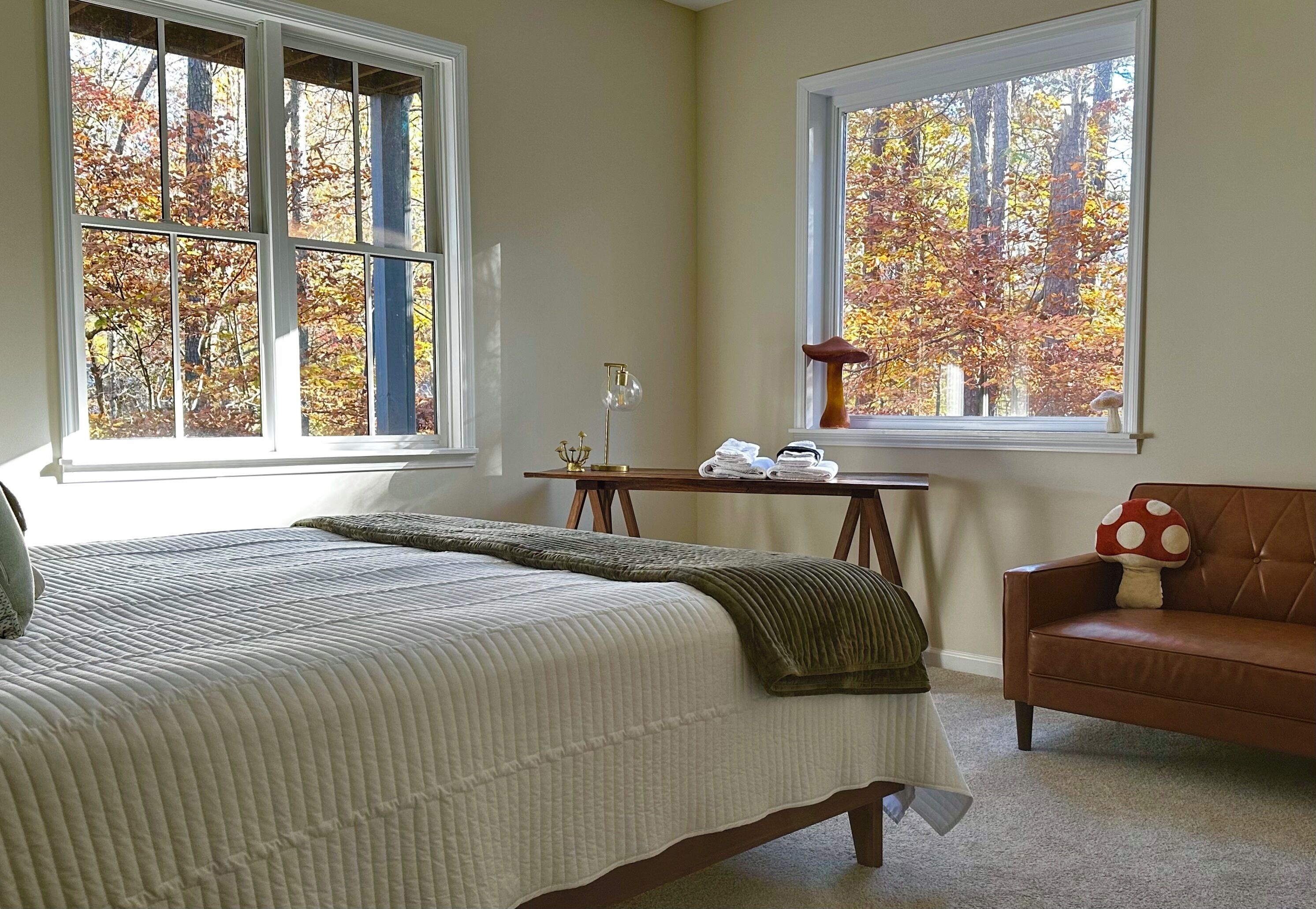 A bedroom with a bed, two windows with autumn foliage views, a wooden desk, and a brown couch.