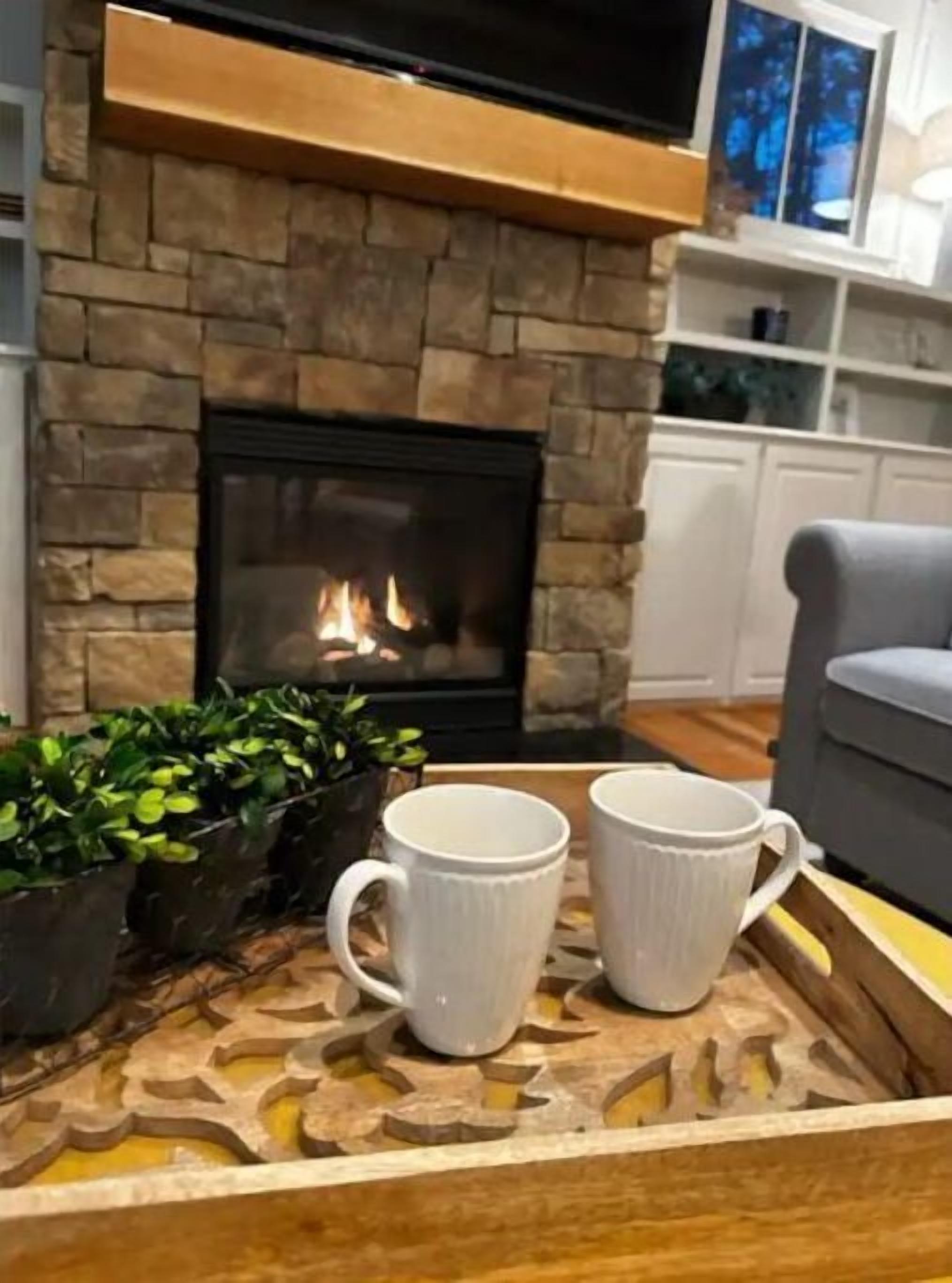 A cozy living room with a stone fireplace, two white mugs on a tray, and a grey armchair.