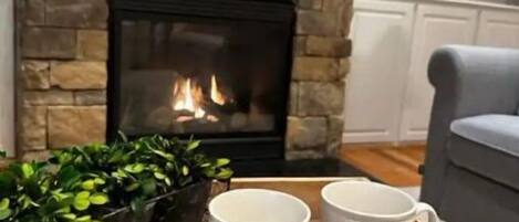 A cozy living room with a stone fireplace, two white mugs on a tray, and a grey armchair.