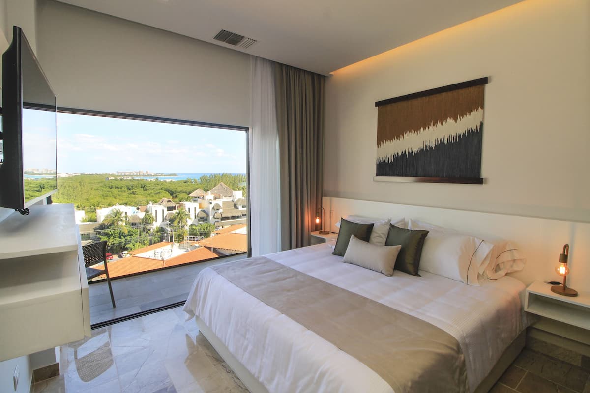 A modern hotel room with a large bed, a flat-screen TV, a city view, and a painting on the wall.