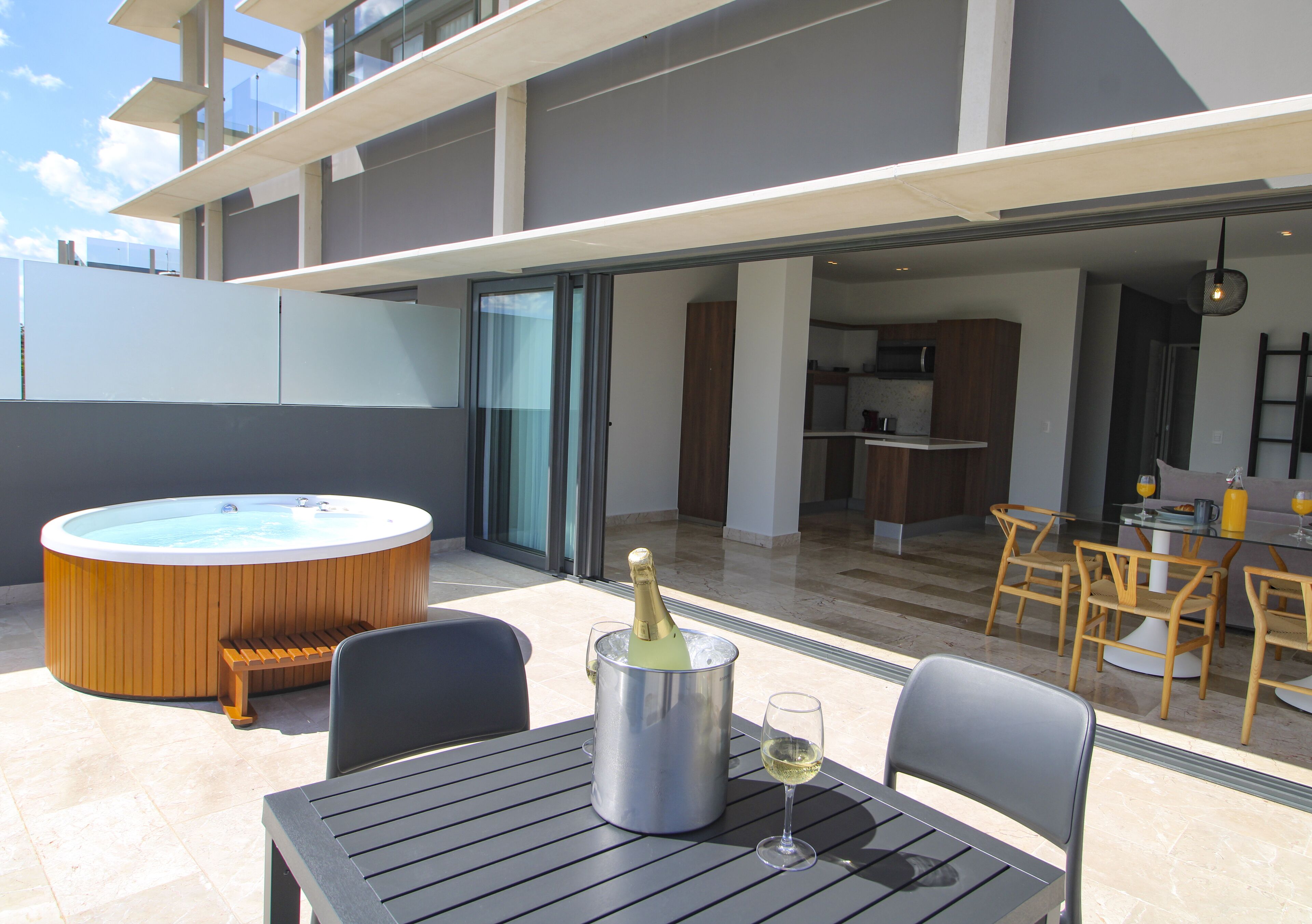 A modern outdoor patio with a hot tub, a table with a bottle of champagne and a glass, and a dining area with chairs and a table.