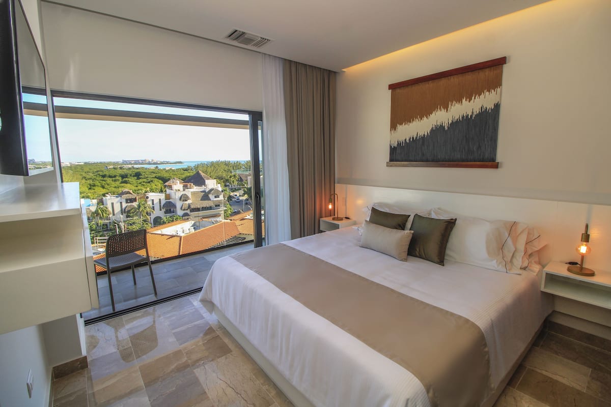 A hotel room with a large bed, a TV, a balcony with a view of the city, and a painting on the wall.
