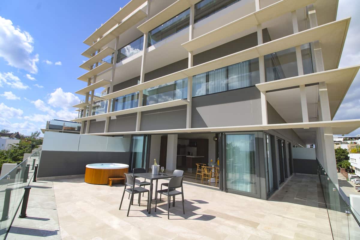 A modern multi-story building with a rooftop patio featuring a table and chairs, a hot tub, and a glass-enclosed balcony.