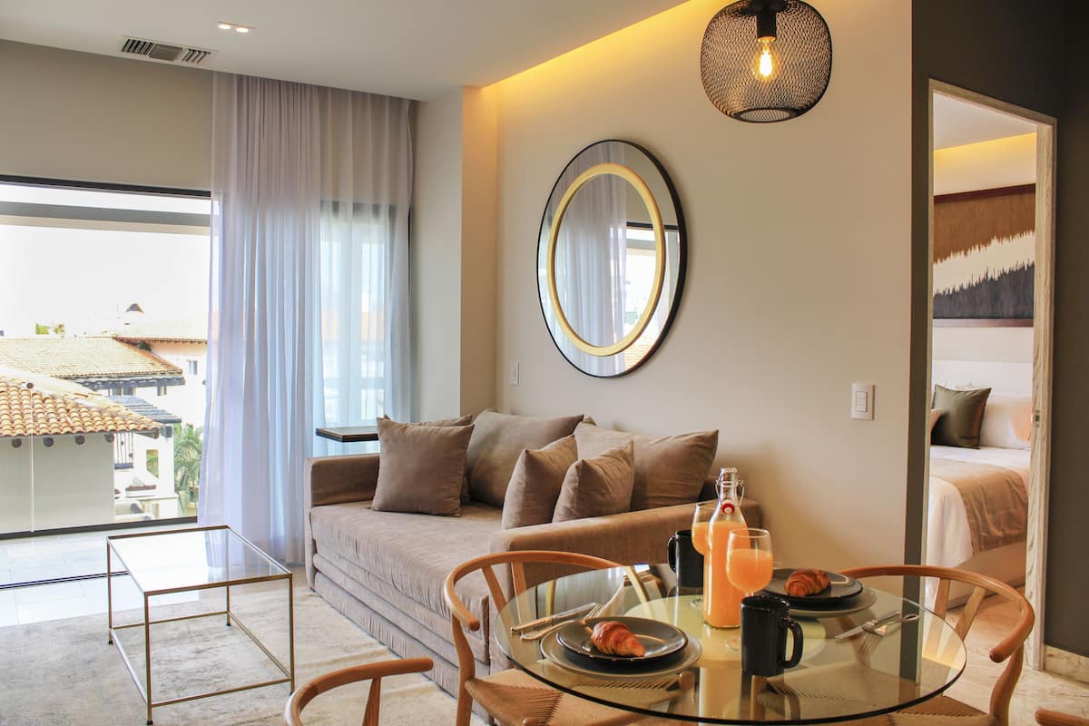 A modern hotel room with a glass dining table, a sofa, and a large window with curtains.