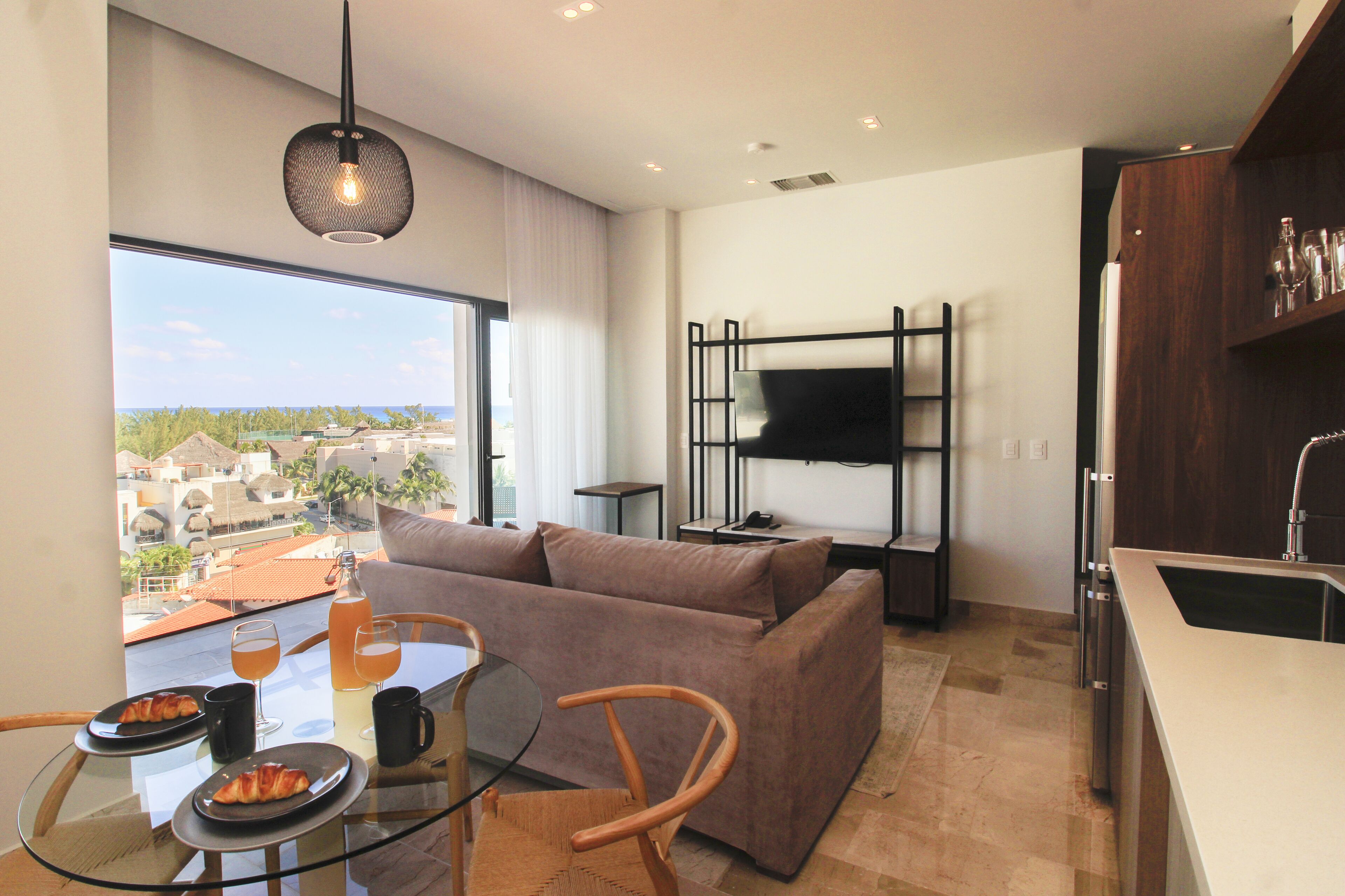 A modern living room with a sofa, a glass table with plates and drinks, a TV mounted on the wall, and a view of the city through large windows.