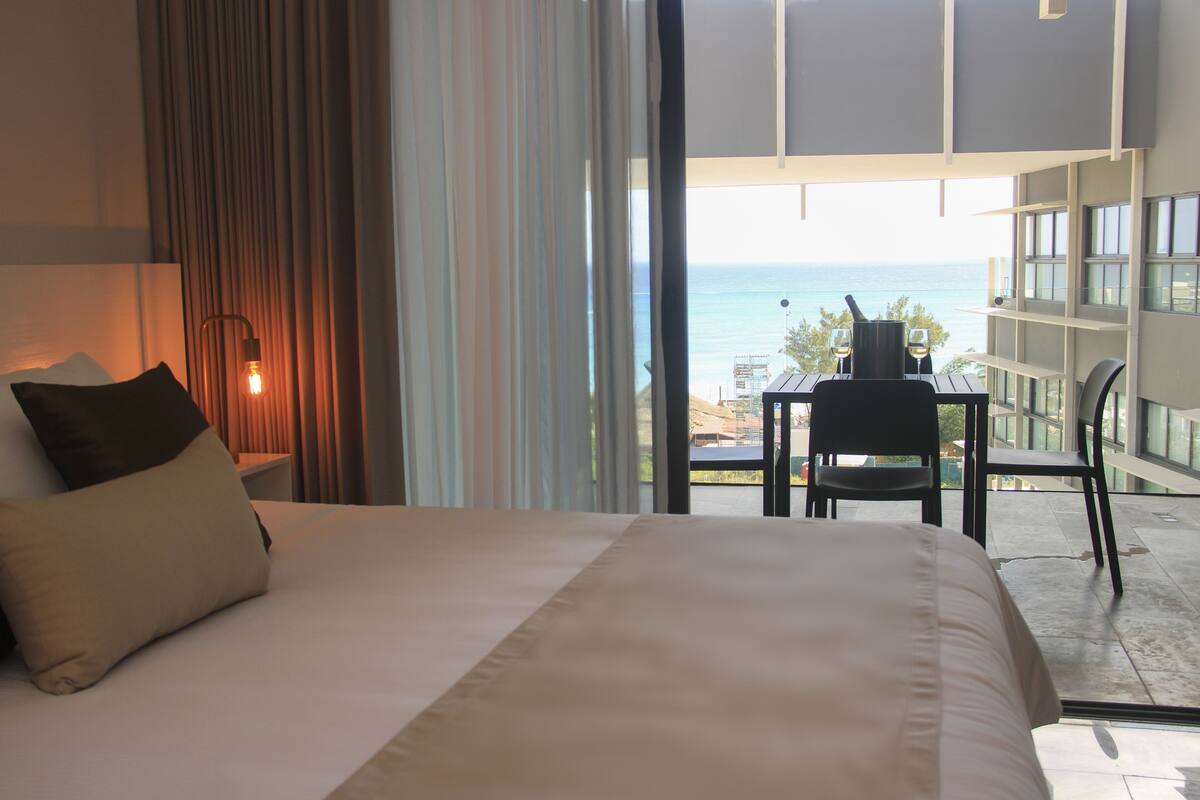 A hotel room with a large bed, a table with chairs, and a view of the ocean.