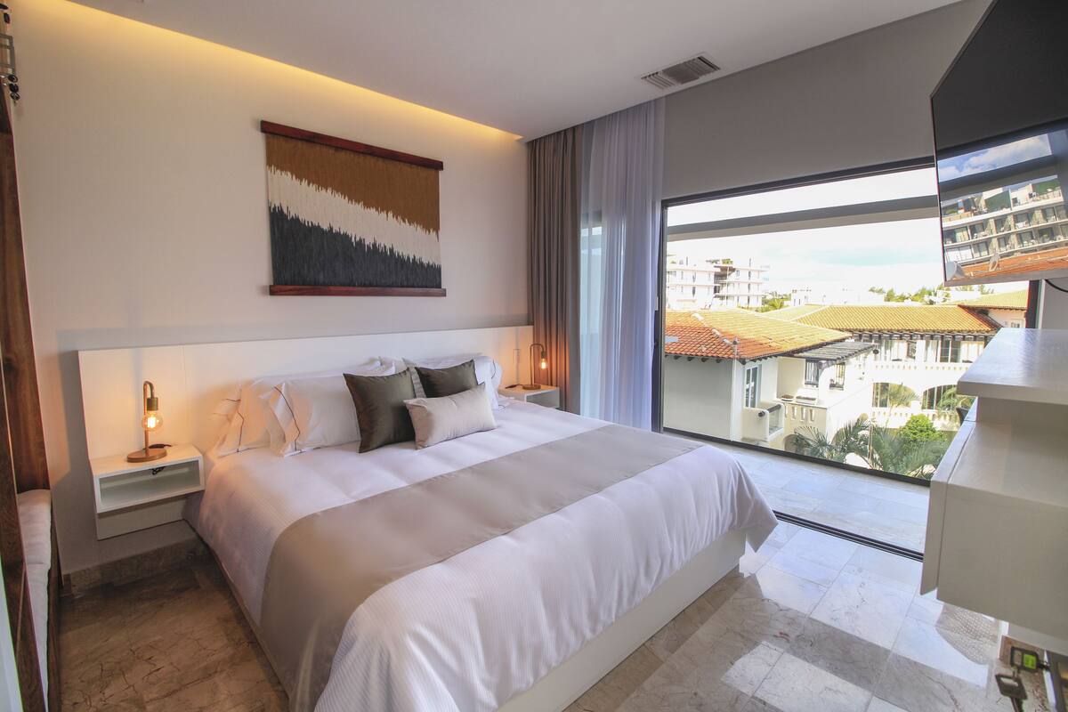 A modern hotel room with a large bed, a nightstand, a wall-mounted painting, and a view of buildings through a large window.
