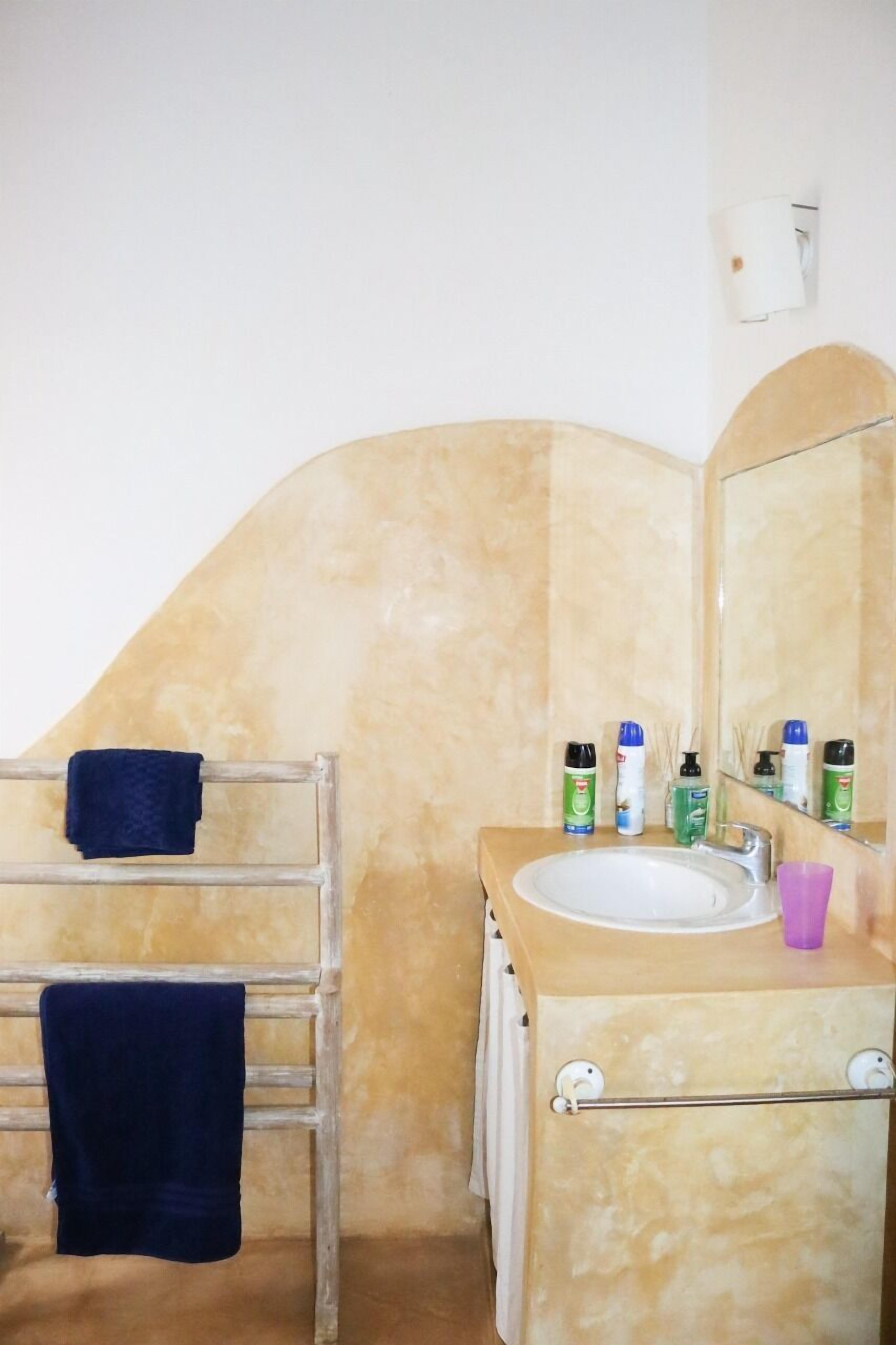 A bathroom with a beige tiled wall, a white sink, and a mirror. There are toiletries on the sink counter.