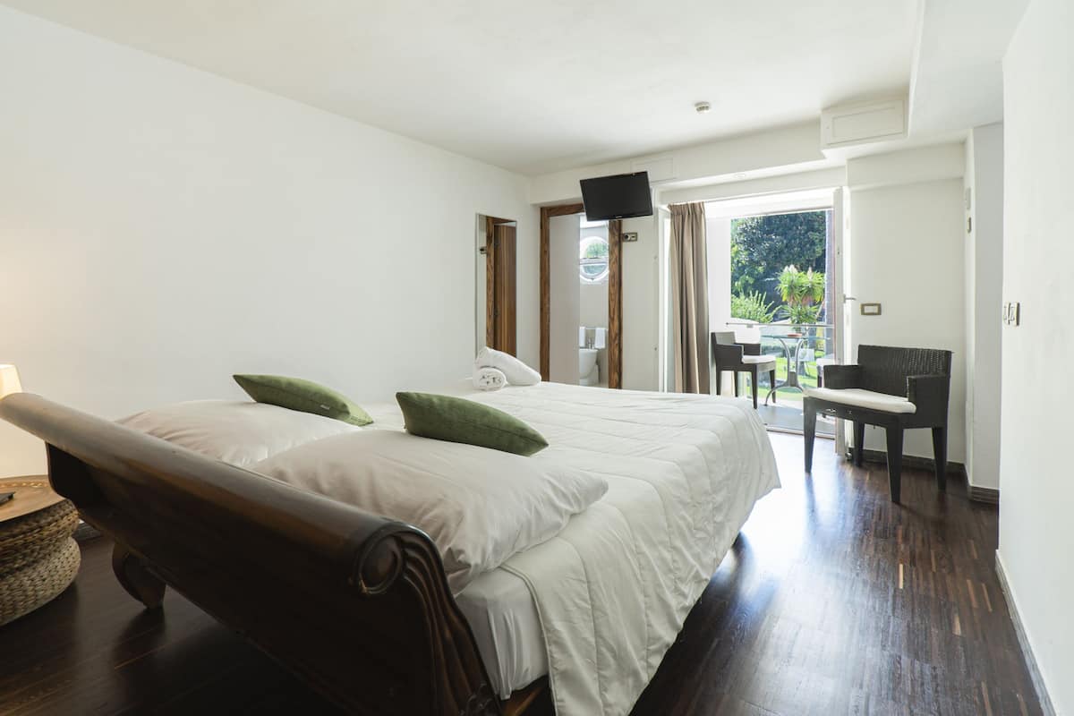 Ein Schlafzimmer mit einem großen Bett, einem Schreibtisch und einem Sessel. Ein Fernseher ist an der Wand montiert und es gibt ein Fenster mit Vorhängen.