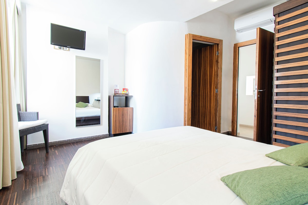 A hotel room with a large bed, a television mounted on the wall, a wooden wardrobe, and a small table with a chair.