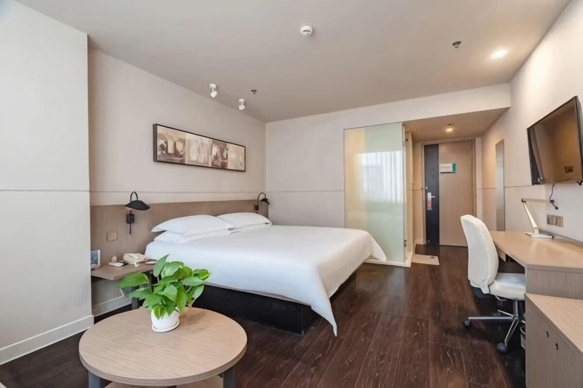 A hotel room with a bed, bedside table, lamp, chair, desk, and a potted plant.