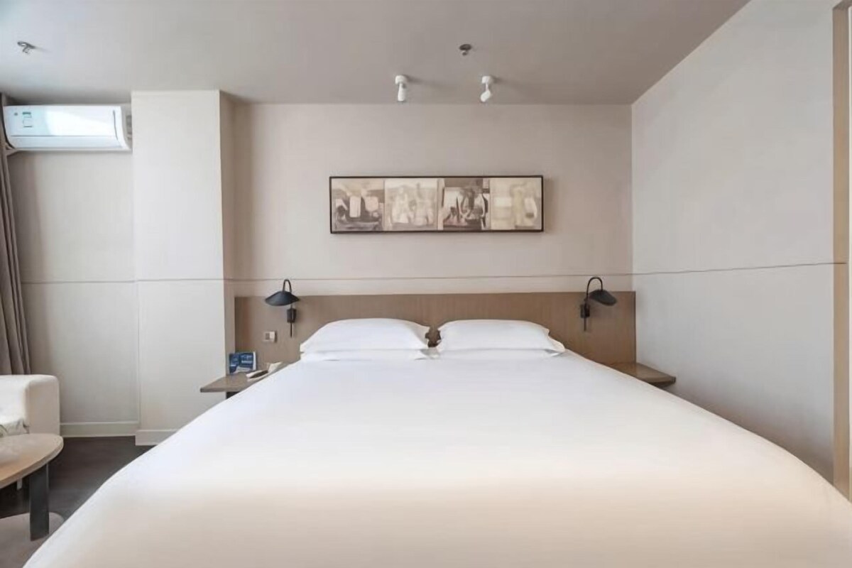 A hotel room with a large bed, bedside tables, a chair, a wall-mounted artwork, and an air conditioning unit.