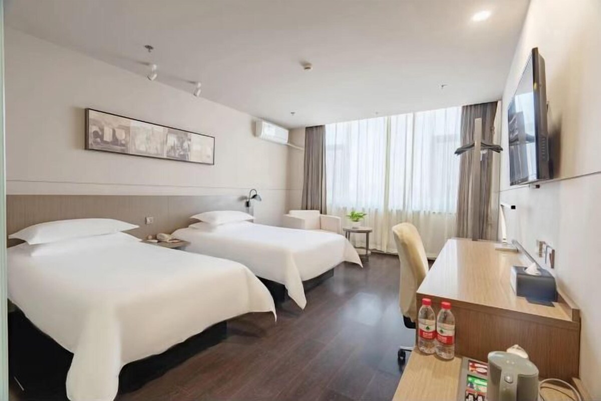 A hotel room with two beds, a desk with a chair, a TV, and a window with curtains.