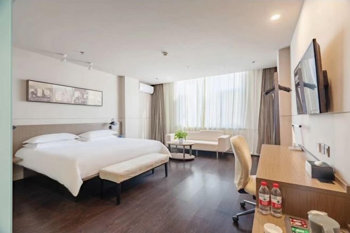 A hotel room with a double bed, a sofa, a desk with a chair, a TV, and a window with curtains.