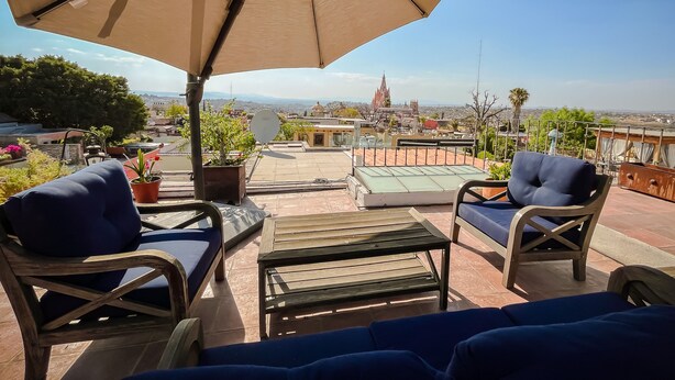 Terraza con mobiliario exterior, una sombrilla grande y vistas a un paisaje urbano distante que incluye una iglesia.